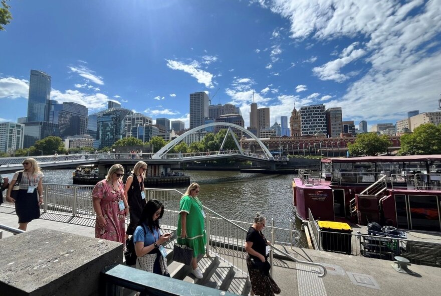 People boarding a cruise boat on the Yarra River with buildings and bridge in background.