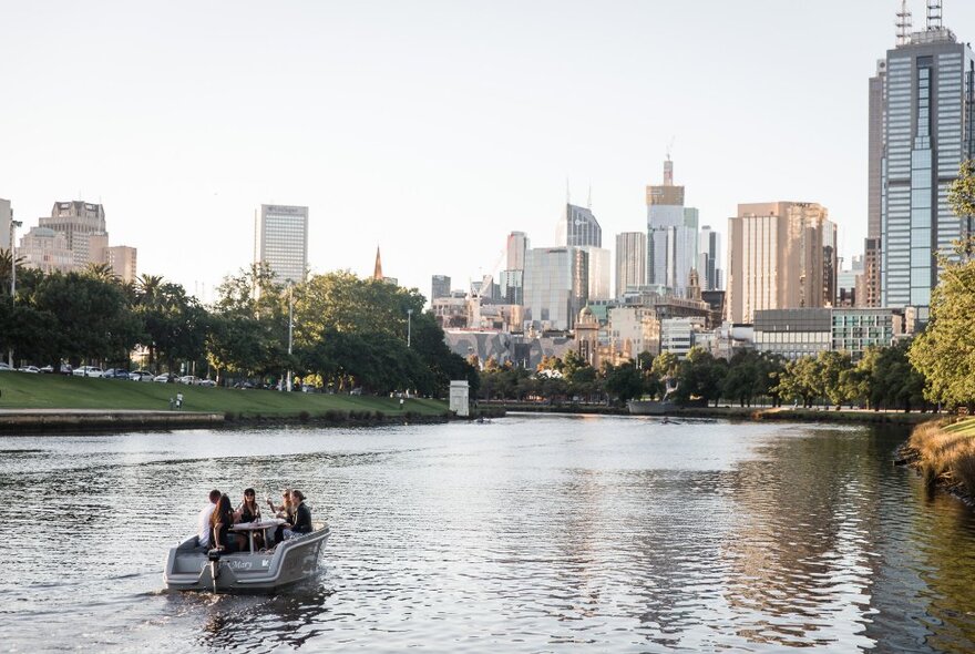 A small boat cruising along the Yarra River in the late afternoon sun, with a view of the Melbourne city skyline in the background.