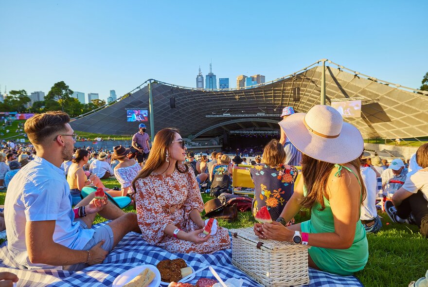 Three people sitting on a picnic blanket with food watching the Sidney Myer Music Bowl stage on a sunny day in the city.