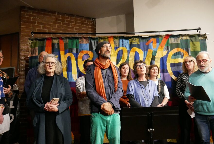 People standing and singing in front of a colourful sign that has the words 'the boite' written on it.