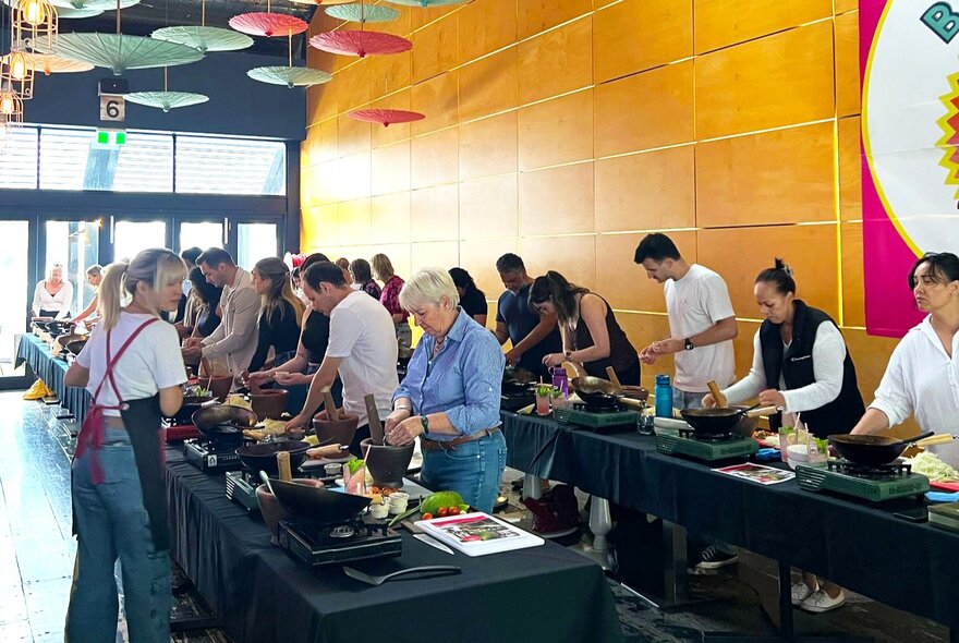 People working at long tables set up with cooking stations and ingredients and cooking utensils during a Thai cooking workshop.