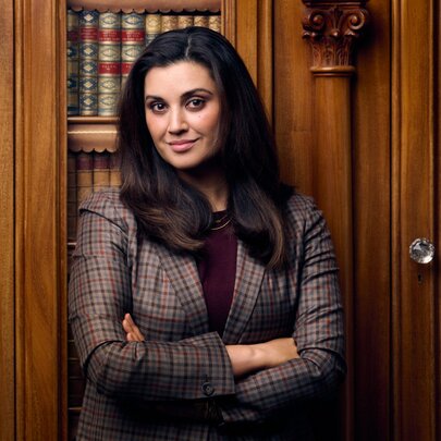 Portrait of a woman standing with arms folded in front of traditional wooden library shelving filled with gold-bound books.