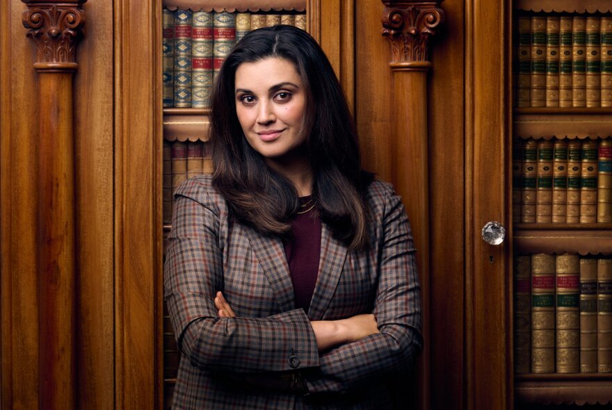 Portrait of a woman standing with arms folded in front of traditional wooden library shelving filled with gold-bound books.