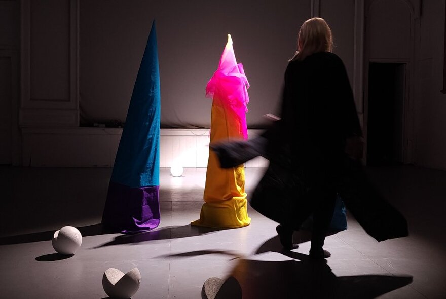 Dark room with a person in the shadows, and two sculptures wrapped in coloured fabric.