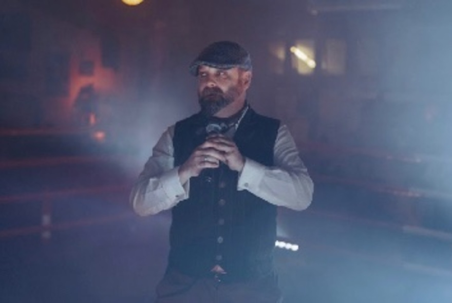 Author Roy Maloy wearing a cap, and a buttoned up waistcoat, holding a microphone in his hands. 