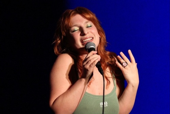 Performer with red hair, wearing a green tank top, singing into a microphone, her eyes closed.