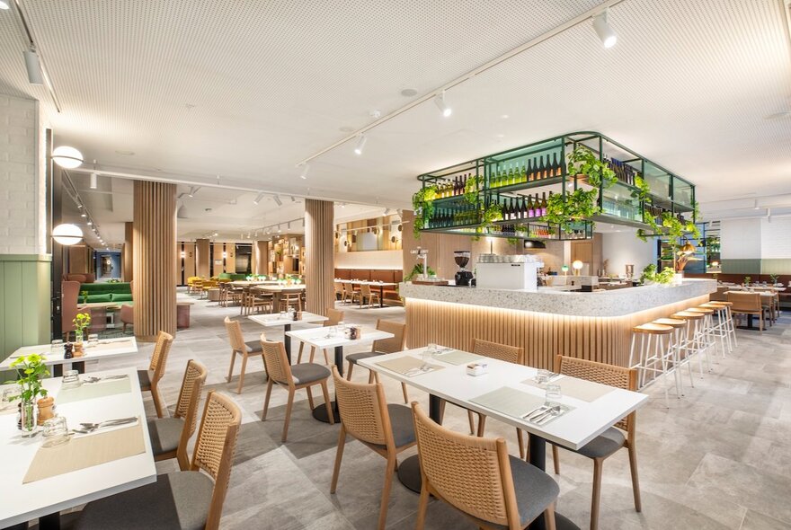 A hotel dining area with blond wood chairs and tables, a central bar area and pops of greenery and plants.