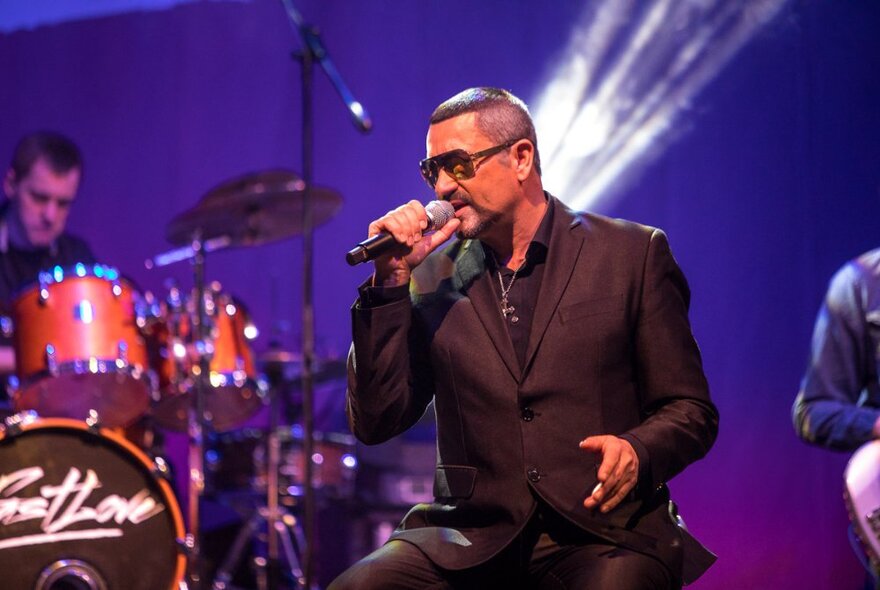 George Michael tribute performer with close-cut hair, sunglasses and suit, singing into microphone on purple-lit stage.