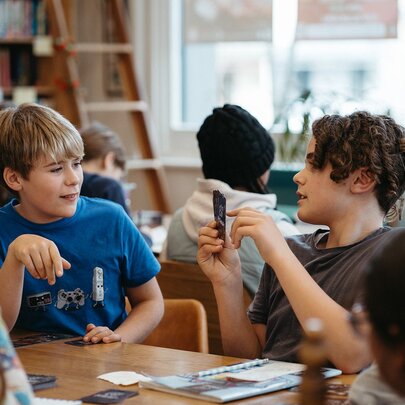 Two teen students writing on notebooks or into laptops and interacting at a table, as part of a creative writing workshop in a studio setting.