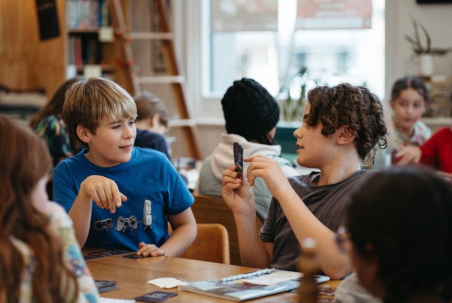 Two teen students writing on notebooks or into laptops and interacting at a table, as part of a creative writing workshop in a studio setting.