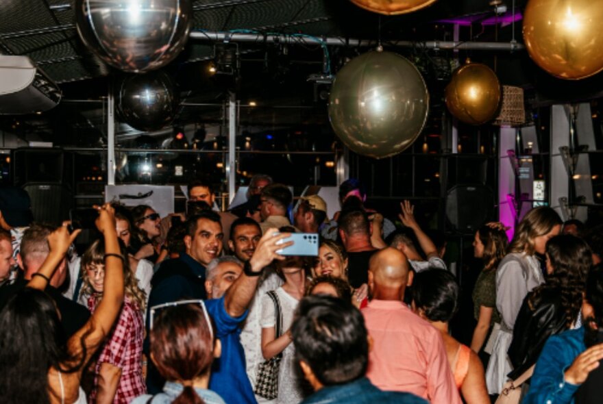 Crowd on a dance floor under mirror balls taking selfies and dancing.