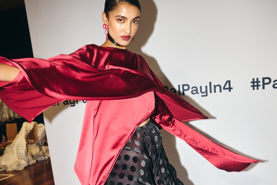 A fashion model posing in a vibrant red, wide-sleeved satin top and a dark polka-dot skirt at a fashion show, against a branding board.