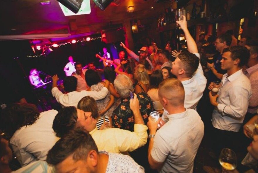 A crowd in a pub enjoying a band on stage, hands in the air. 