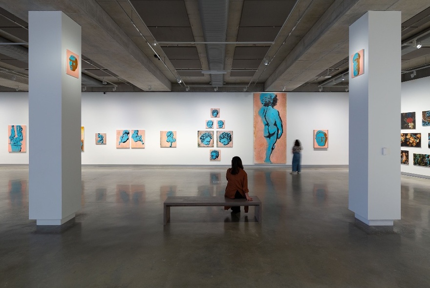 Person seated on a central bench in a large gallery space with white pillars and blue artworks on a long white wall.