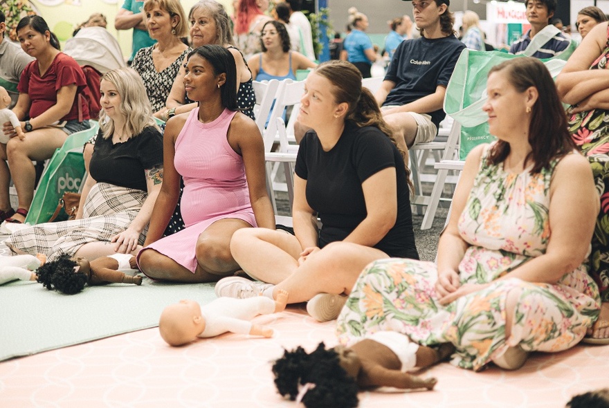 Pregnant mums seated on the floor with baby dolls lying in front of them, watching something out of frame.