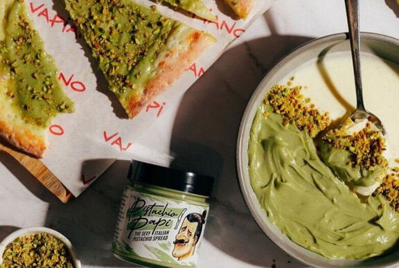 Slices of pistachio pizza and pistachio panna cotta on a table, with a jar of pistachio spread.