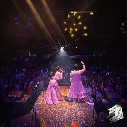Two Bhajan performers in glittering traditional clothes singing on stage, seen from behind with the audience in front of them.