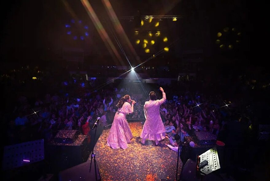 Two Bhajan performers in glittering traditional clothes singing on stage, seen from behind with the audience in front of them.