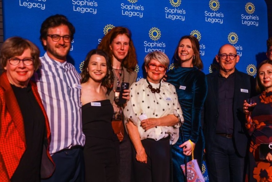 A group of smiling people standing in front of a branded blue backdrop for Sophie's Legacy, an organization that advocates for the wellbeing and mental health of veterinary professionals.