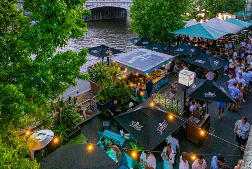 People standing on the wharf outside Riverland Bar at twilight, the Yarra river in the background, the area set up with large umbrellas. 