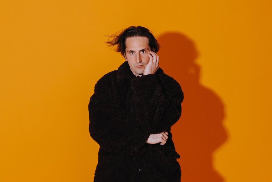 Comedian Luke McQueen wearing a heavy dark coat, with one arm crossed in front of his chest and the other one hand placed near his face, standing in front of a bright orange backdrop.