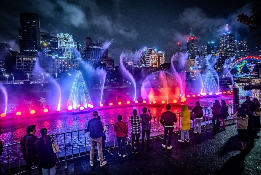 Rainbow lights on water jets and a giant bauble on the river at night.