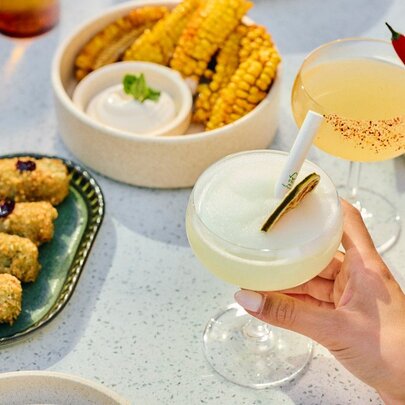 Two cocktails and share plates of Mexican food on a white table.