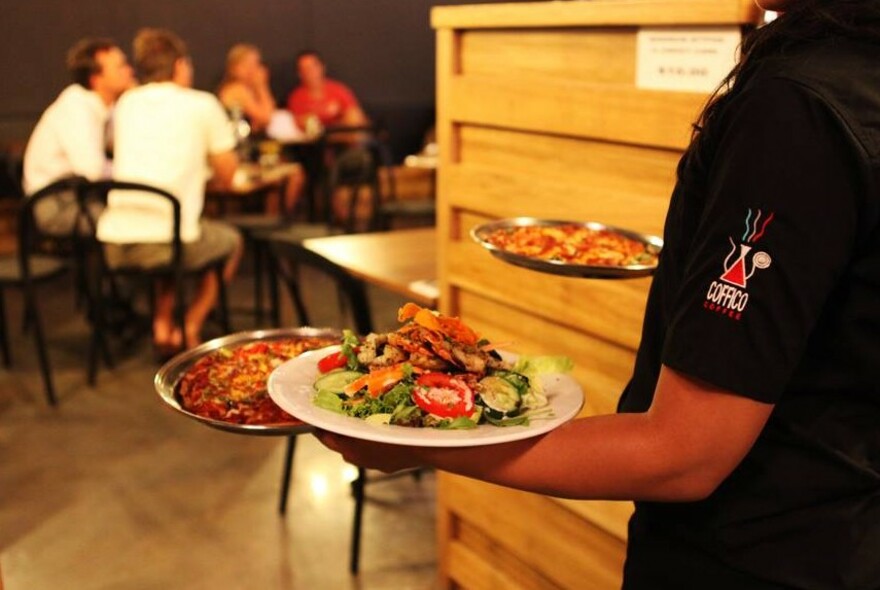 Waiter carrying two pizzas and dish of meat and salad to customers sitting at tables.