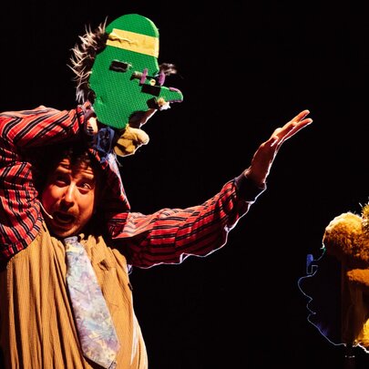 A puppeteer on stage with a green puppet made from recycled and found materials.