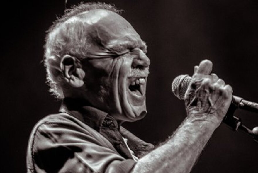 Guitarist Dave Hole singing into a microphone with his eyes closed.