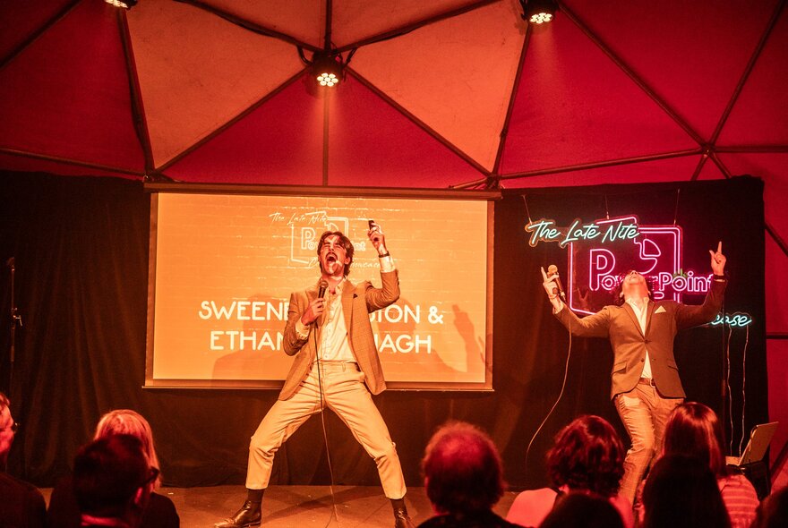 Two performers on a small stage, both wearing suits, behind them is a screen with a projection on it.