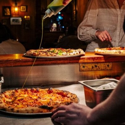 Pizzas on a restaurant pass, someone drizzling olive oil over the top. 