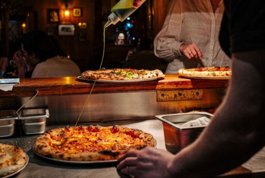 Pizzas on a restaurant pass, someone drizzling olive oil over the top. 