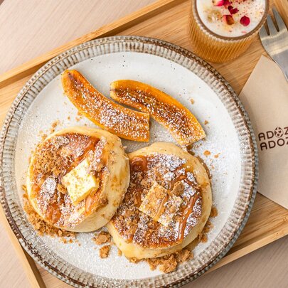 Souffle-style fluffy pancakes with caramelised banana and caramel sauce on a round plate alongside a fork, napkin and drink on a wooden tray.