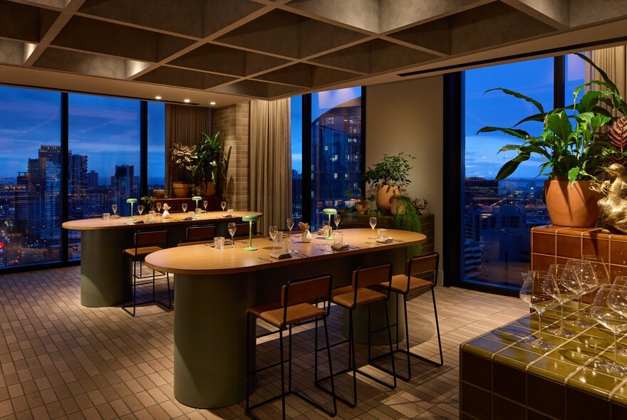 A modern, dimly lit restaurant dining room at dusk with floor-to-ceiling windows showing a blue city skyline.