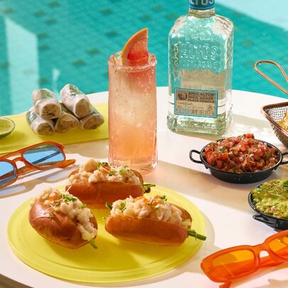 A table of snacks next to a pool with a prawn roll, avocado and fried tofu rice paper rolls, corn chips with guacamole and pico de gallo, and a paloma cocktail with a bottle of tequila.