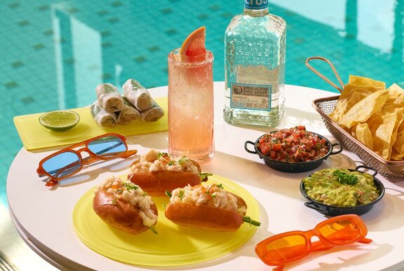 A table of snacks next to a pool with a prawn roll, avocado and fried tofu rice paper rolls, corn chips with guacamole and pico de gallo, and a paloma cocktail with a bottle of tequila.