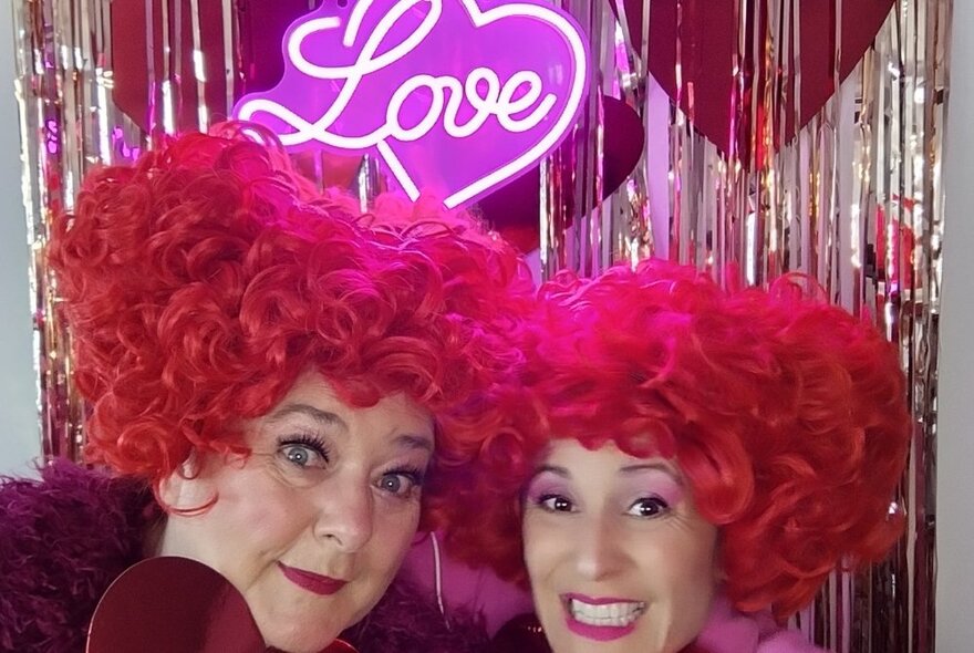 Two women wearing curly red wigs posing beneath a pink neon sign with the word 'love' and silver glitter curtain.