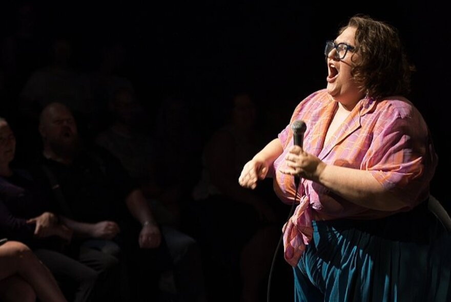 Large comedian performing on stage, holding a microphone with mouth wide open.