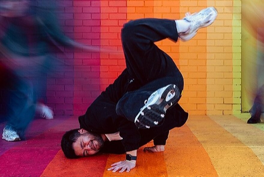 A breakdancer dancing on a rainbow painted floor, in the middle of a spinning move with his head on the floor.
