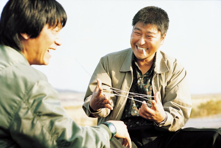 A still image from the South Korean film Memories of Murder showing two men enjoying each other's company, one holding a cat's cradle in his hands.