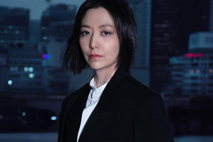 A woman, Ayoung Kim, with a black bob haircut, wearing a dark jacket over a white shirt, looking directly at the viewer, in the twilight background tall city buildings are visible..