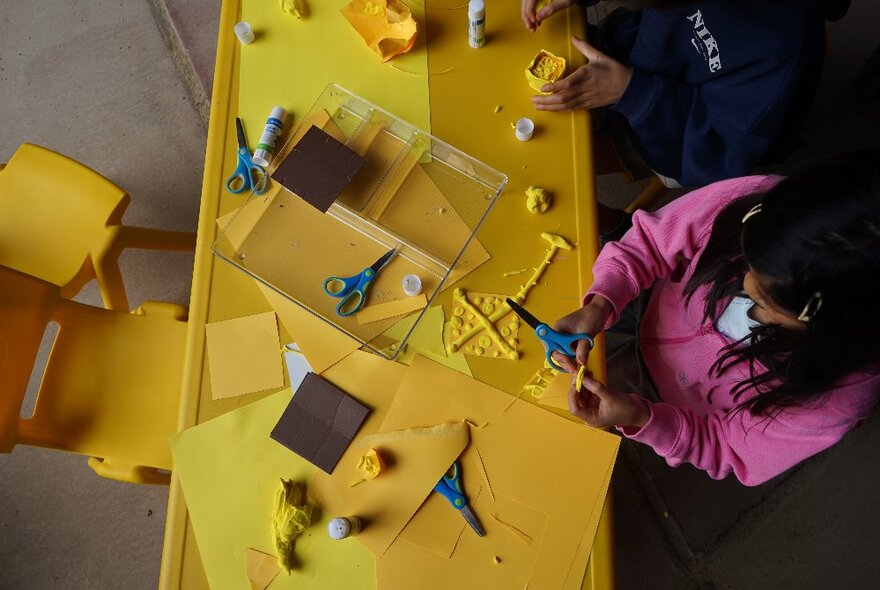 Kids working at a yellow craft table with yellow crafting materials and blue-handled scissors on it, seen from above.