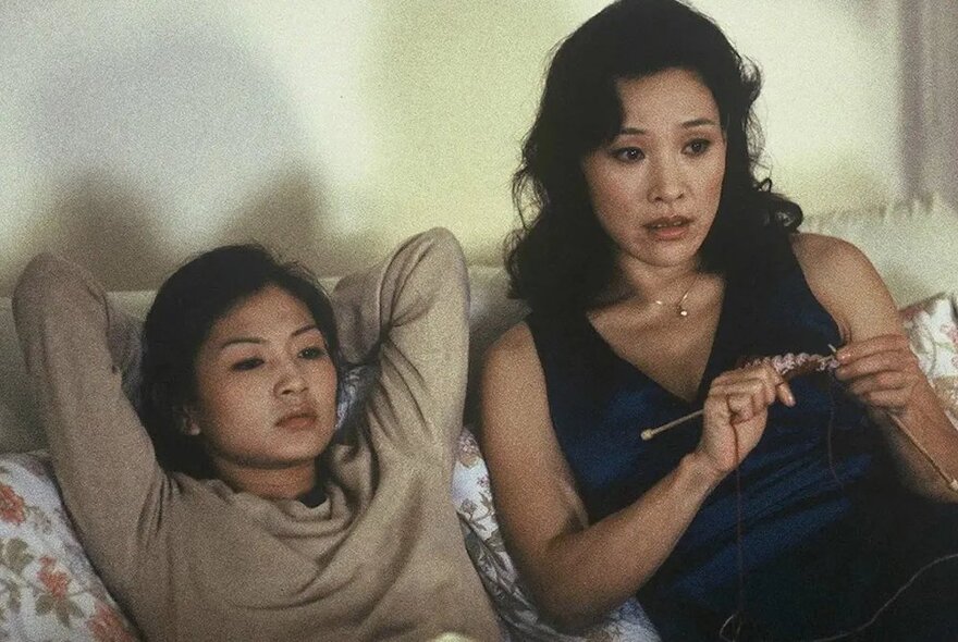 Film still of two women in a relaxed pose on a couch, looking straight ahead, one of them knits.