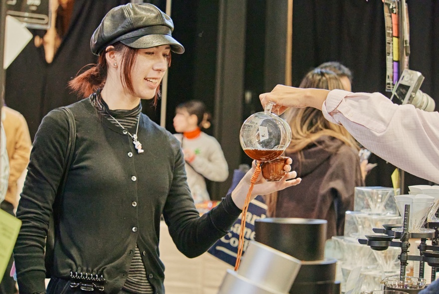Person holding a cup while another person fills it with coffee from a glass jug in a market setting.