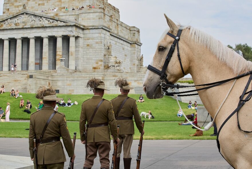 Rear views of three men in army uniforms standing to attention with rifles by their side, next to a camel coloured horse, the Shrine of Remembrance building in the background.