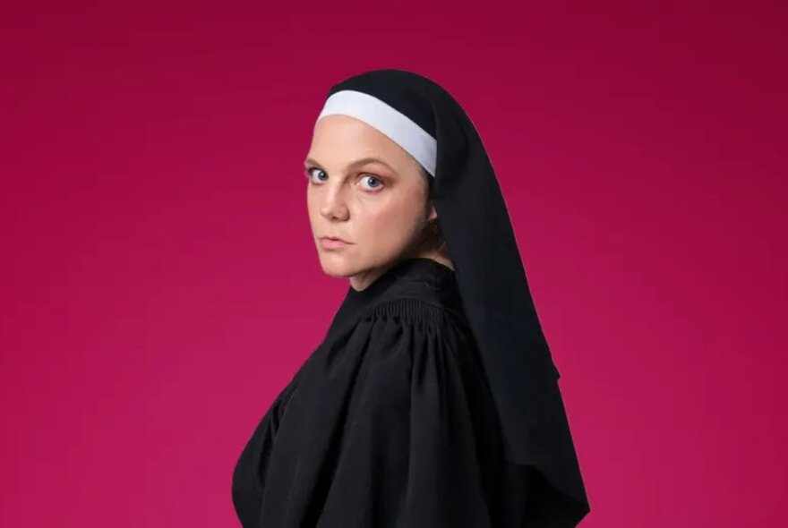 Performer Claire Robin wearing a nun's costume and habit, posed against a magenta background.