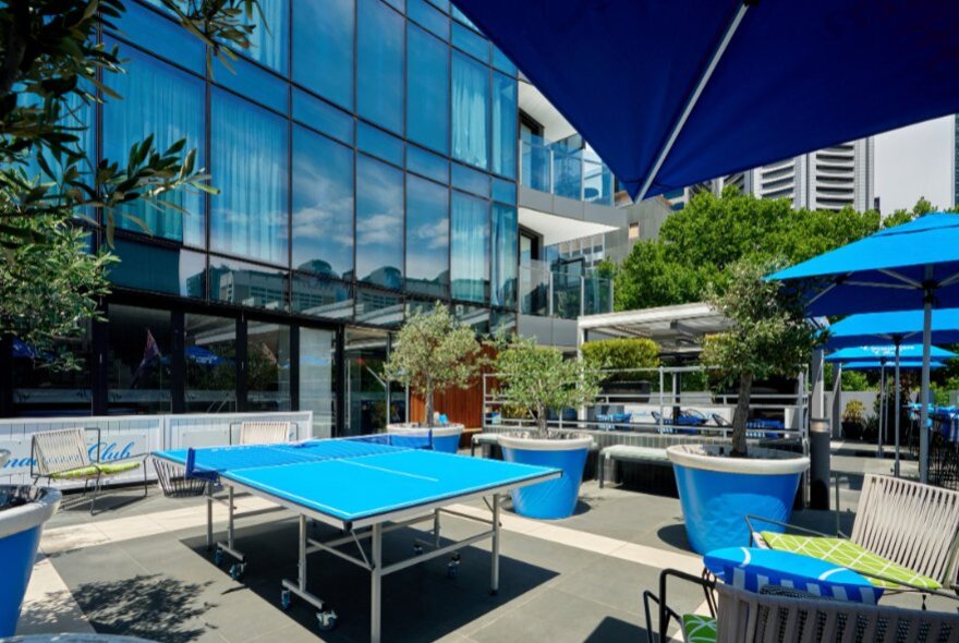 An outdoor terrace area with a blue ping-pong table and blue patio umbrellas next to a modern glass-facade building.
