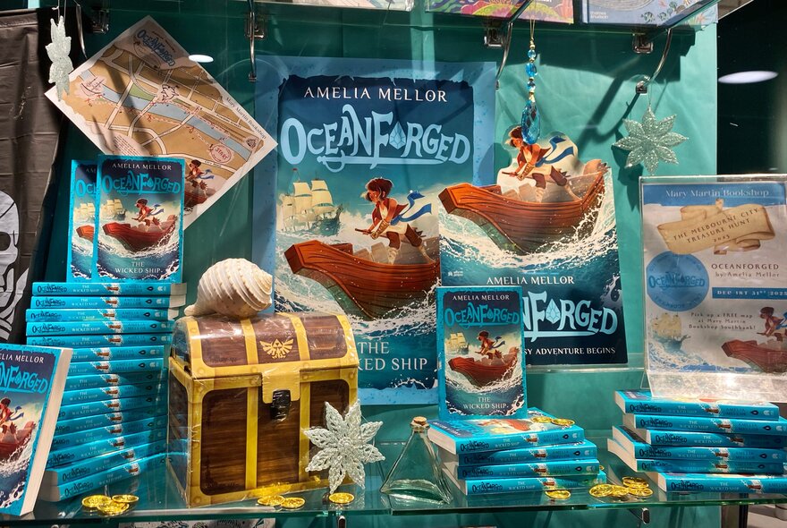 Piles of Amelia Mellor's book, Oceanforged, displayed with a treasure chest, treasure maps and posters.