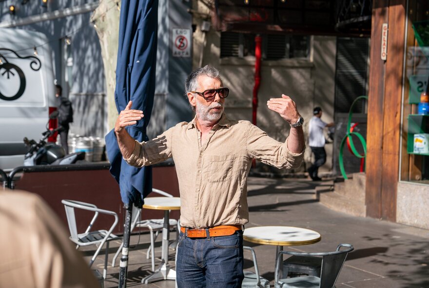 A tour leader wearing sunglasses gesticulating in front of cafe chairs and tables. 
